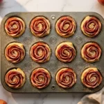 Twelve elegant apple roses made with puff pastry and thinly sliced red apples in a muffin tin dusted with powdered sugar