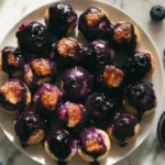 Baked blueberry fritter bites on a white plate with shiny caramelized blueberry glaze drizzled down the sides